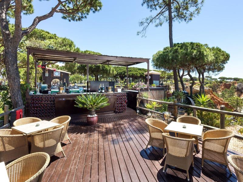 Terrasse mit Holzboden, mehreren Tischen und Stühlen, umgeben von Bäumen und Pflanzen.