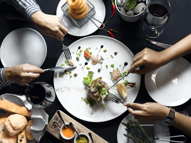 Mehrere Personen greifen gemeinsam auf einem dunklen Tisch zu, in der Mitte ein Teller mit Resten.