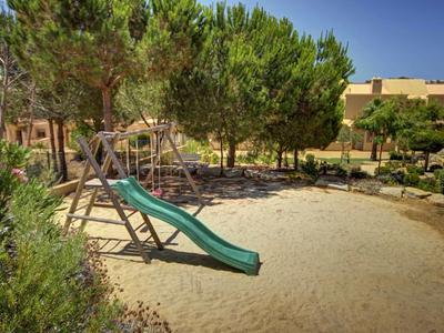 Spielplatz mit Rutsche, umgeben von Bäumen und Gebäude im Hintergrund.