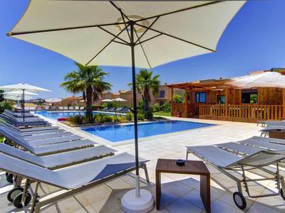 Poolbereich mit Liegestühlen, Sonnenschirmen, Palmen und Holzhütten bei klarem Himmel.