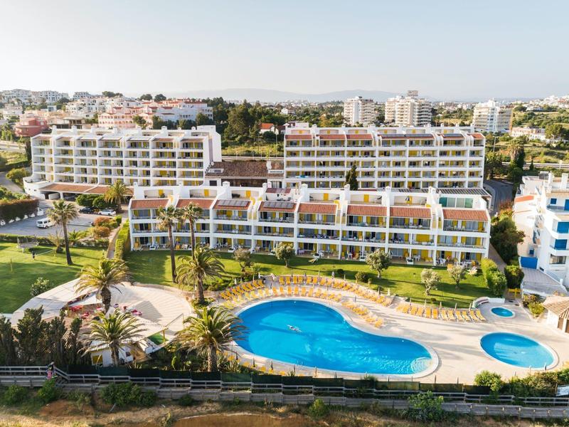 Hotelcomplex met grote zwembaden, parasols en nabijgelegen woongebouwen onder een heldere lucht.