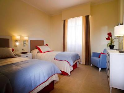 Hell eingerichtetes Hotelzimmer mit zwei Einzelbetten und einem blauen Sessel am Fenster.