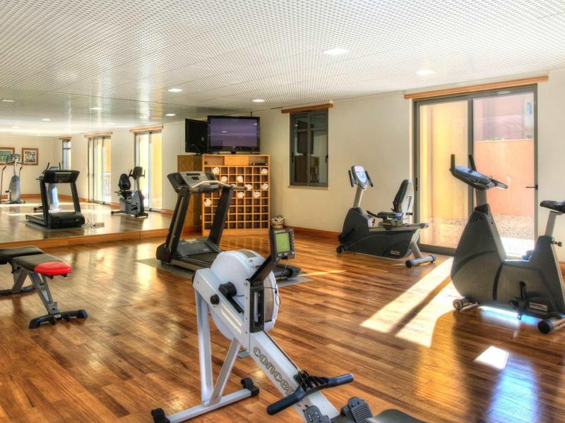 Salle de sport lumineuse avec sol en bois, divers appareils d'exercice et grandes fenêtres.