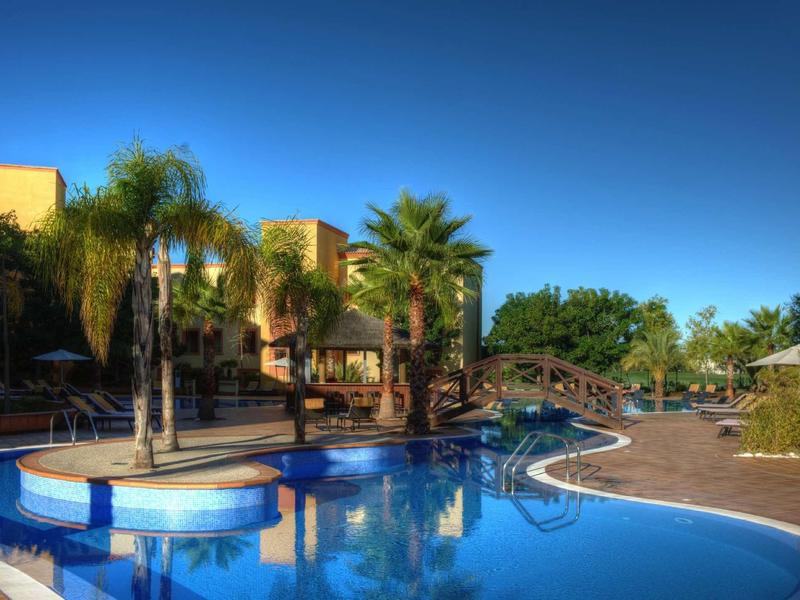 Piscine d'hôtel avec palmiers, chaises longues et ciel bleu clair par une journée ensoleillée.