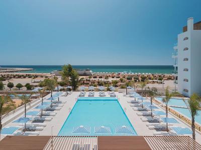 Moderner Außenpool mit Liegestühlen, Palmen und Meer am Horizont bei klarem Himmel.