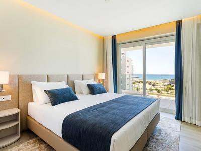 Helles Schlafzimmer mit großem Bett, blauen Akzenten und Blick auf das Meer durch eine Glaswand.