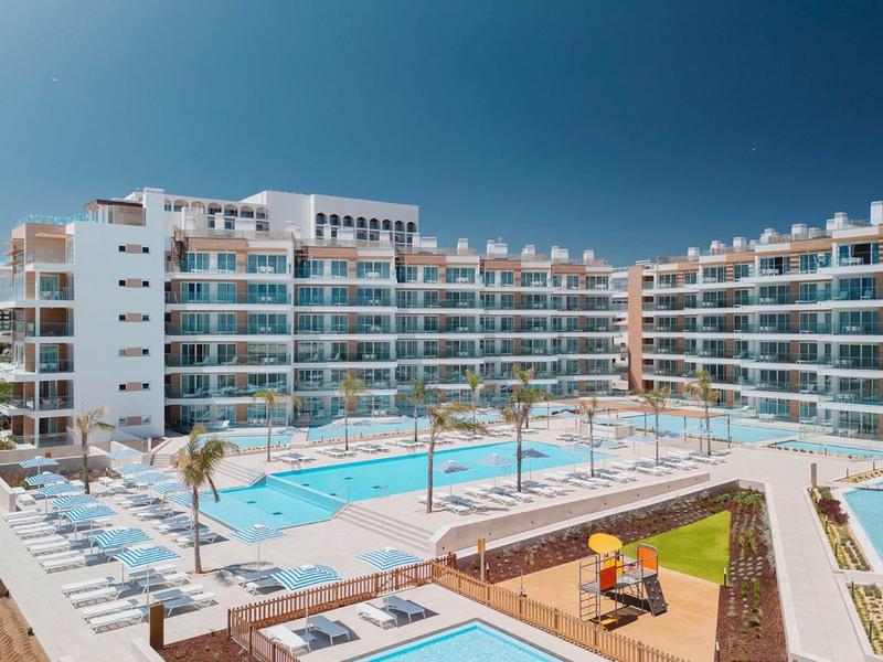 Große Wohnanlage mit Pool, Liegestühlen und Palmen unter klarem, blauem Himmel.