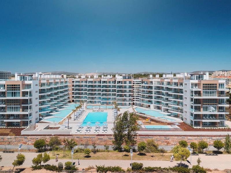 Große Wohnanlage mit mehreren Stockwerken und zwei Pools vor klarem blauem Himmel.