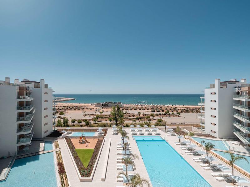 Moderner Hotelpool mit Sonnenschirmen zwischen zwei weißen Gebäuden, Meer und Strand im Hintergrund.