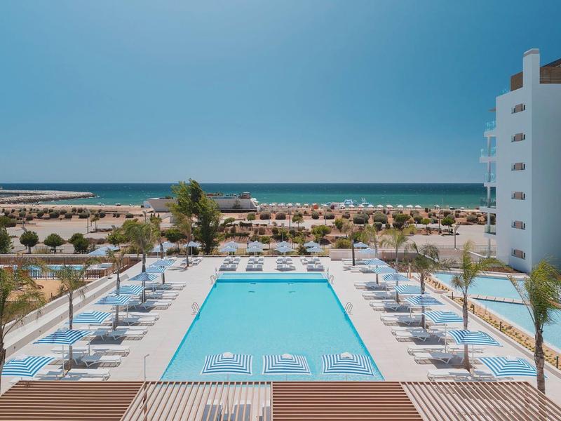 Moderner Außenpool mit Liegestühlen, Palmen und Meer am Horizont bei klarem Himmel.