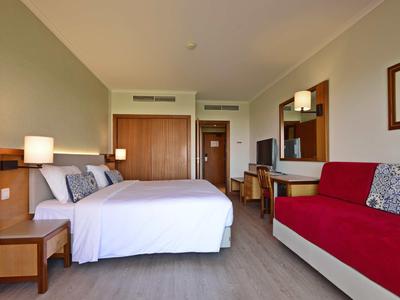 Jasno urządzony pokój hotelowy z dużym łóżkiem, czerwoną sofą i drewnianymi meblami.