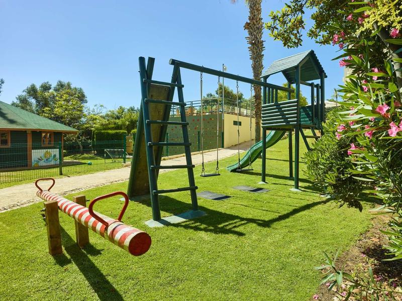 Speeltuin met schommel, glijbaan en wip op groen gras onder een blauwe lucht.