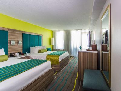 Hotelzimmer mit zwei Doppelbetten, gelben und grünen Akzenten und einem großen Fenster mit Vorhängen.