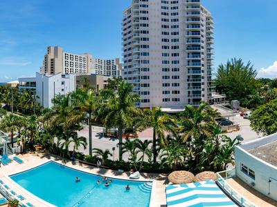 Großer Hotelpool mit Palmen umgeben, Sonnenliegen und hohen Wohngebäuden unter blauem Himmel.