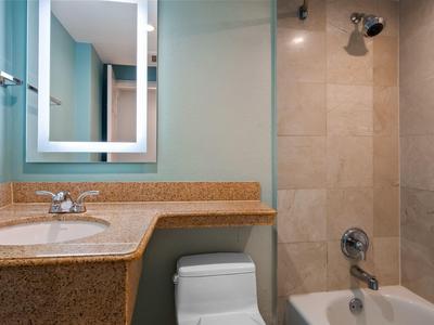 Modernes Badezimmer mit beigen Fliesen, Granitwaschbecken, beleuchtetem Spiegel und Badewanne mit Dusche.