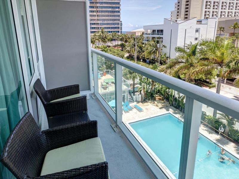 Balkon mit zwei Stühlen und Glasgeländer, Blick auf blauen Pool und Stadtgebäude im Hintergrund.
