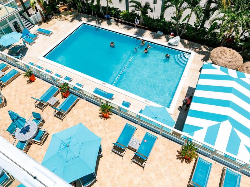 Großes blaues Schwimmbad mit Liegestühlen, Sonnenschirmen und Palmen, sommerliche Atmosphäre.