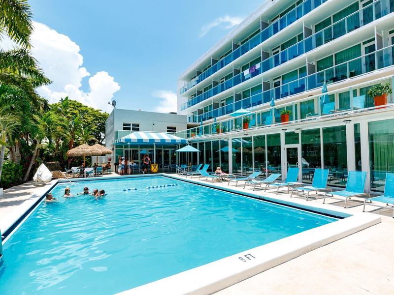 Moderner Außenpool neben Hotel mit Liegestühlen, Palmen und blauem Himmel im Hintergrund.