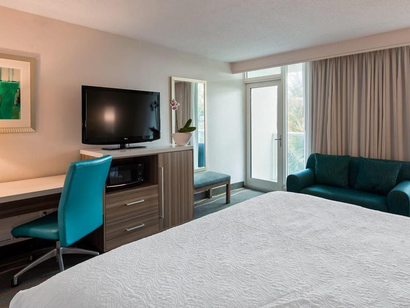 Modernes Hotelzimmer mit Doppelbett, Schreibtisch, Fernseher und Sofa, helle Fenster mit Vorhängen.