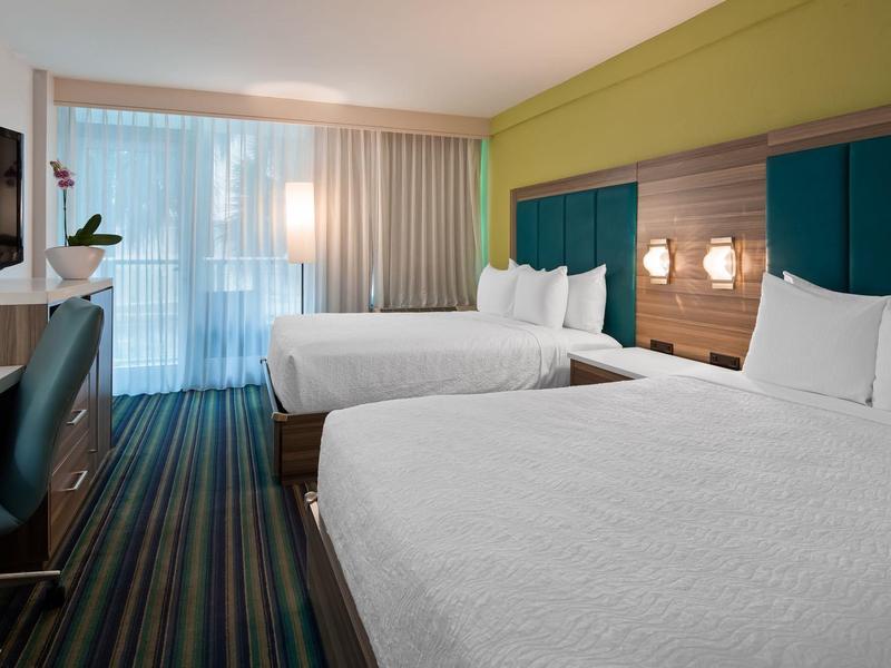 Modernes Hotelzimmer mit zwei großen Betten, blau-grünen Kopfteilpaneelen und gestreiftem Teppichboden.