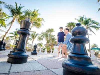 Kinder spielen Schach im Freien auf einem übergroßen Schachbrett mit Palmen im Hintergrund.