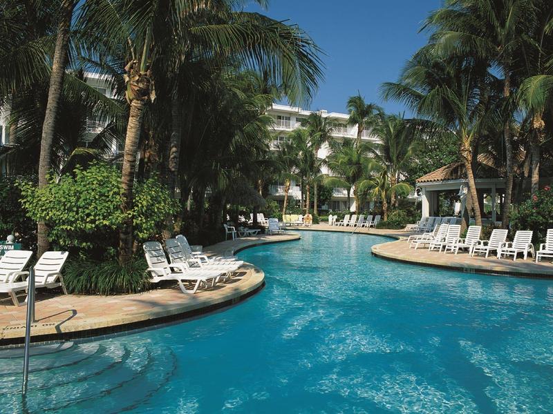 Large pool surrounded by lounge chairs and palm trees in front of a white hotel building.