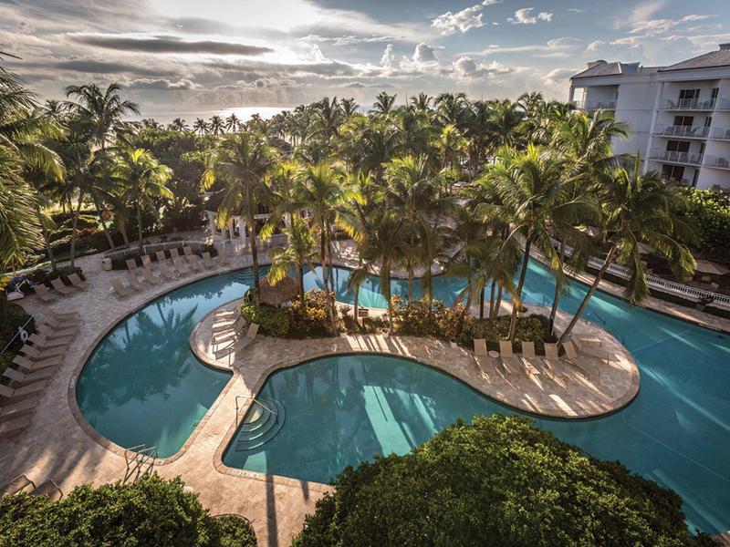 Large hotel pool with palm trees and lounge chairs in tropical setting at sunset.