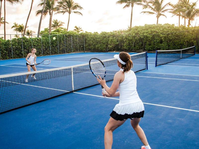 Two people play tennis on a court with palm trees in the background at sunset.