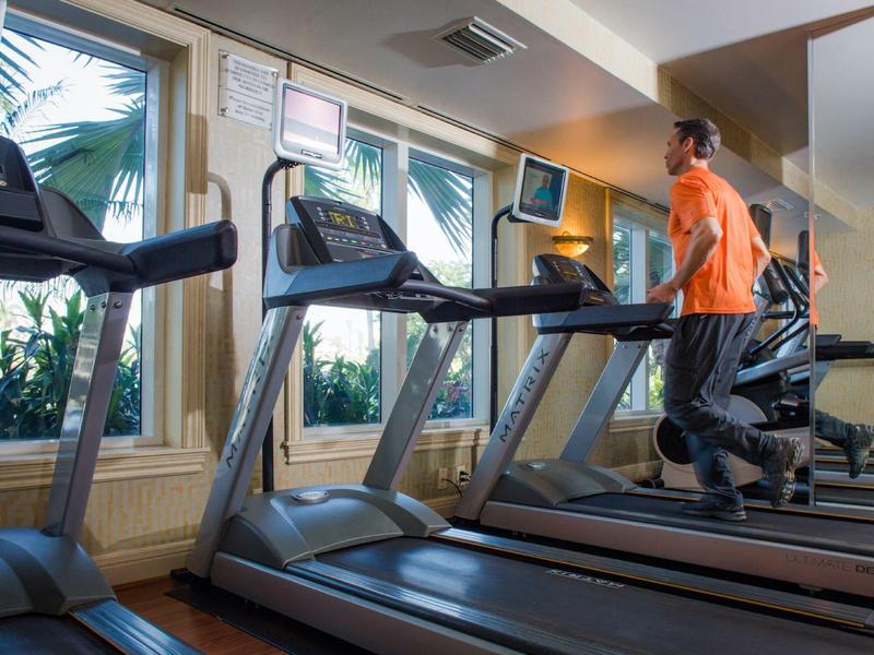 Man running on a treadmill in a bright gym with large windows.