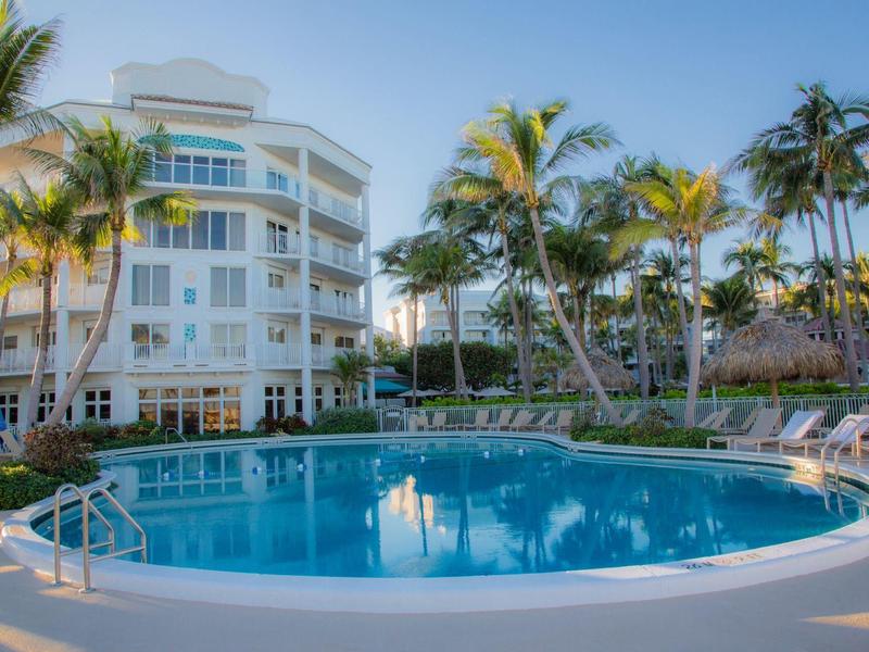Round swimming pool in front of white multi-story hotel with palm trees in clear weather.