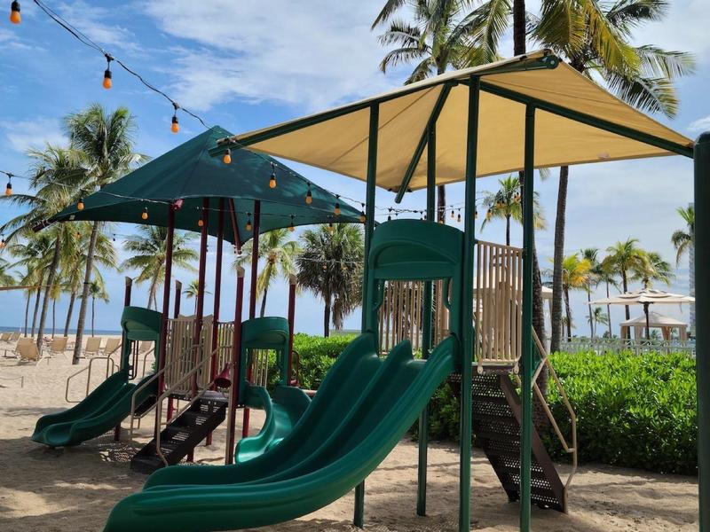 Children's playground with green slides and sunshade at the beach under palm trees.
