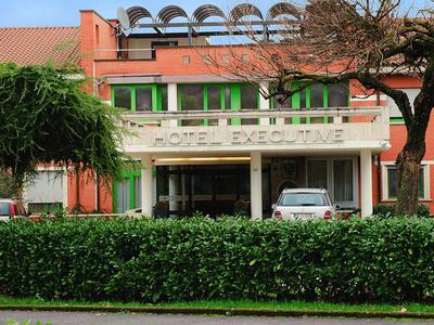 Rotes Backsteingebäude mit grün-gerahmten Fenstern, vor ihm Autos und dichte Hecke, blauer Himmel.