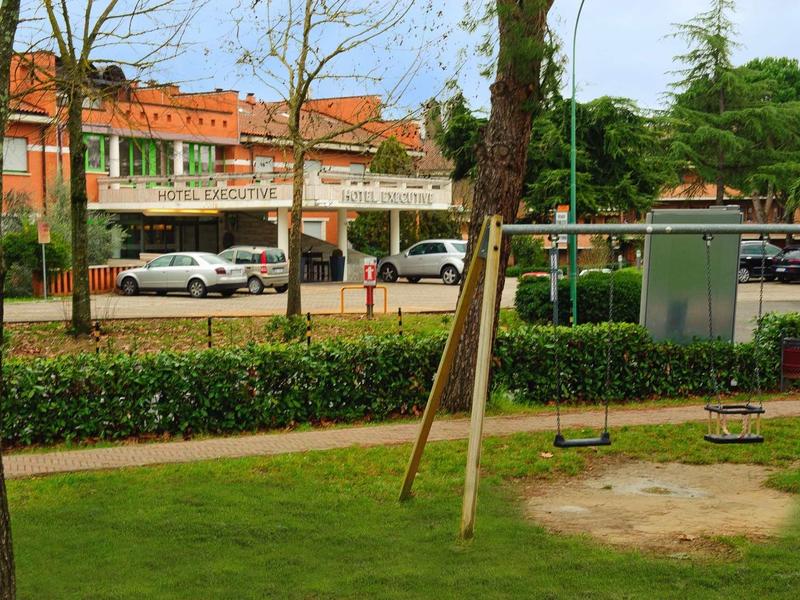 Ein leerer Spielplatz mit zwei Schaukeln, umgeben von Bäumen und Gebäuden im Hintergrund.