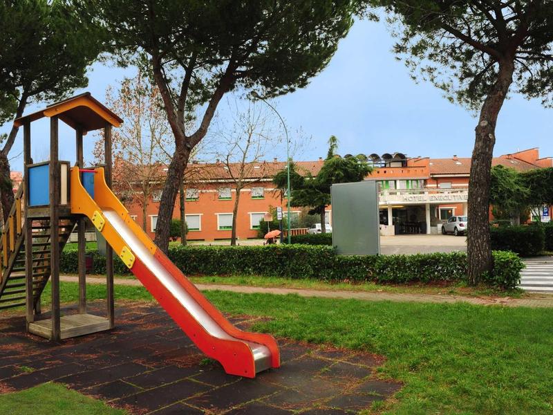 Ein bunter Spielplatz mit Rutsche, umgeben von grünen Bäumen und Gras vor Häusern.