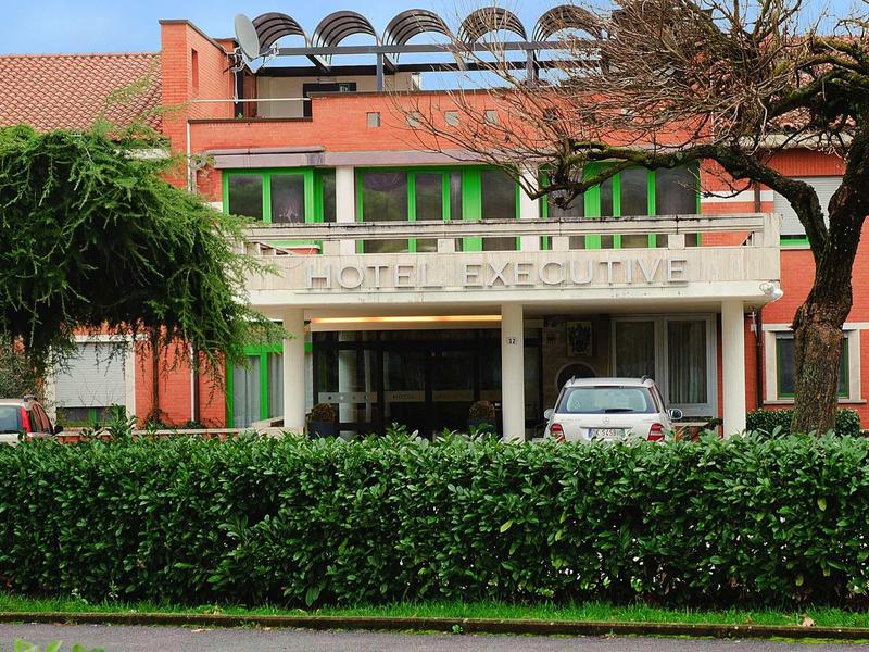 Rotes Backsteingebäude mit grün-gerahmten Fenstern, vor ihm Autos und dichte Hecke, blauer Himmel.