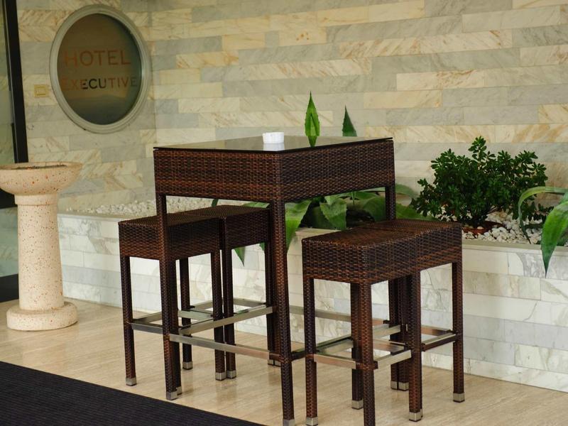 Dunkle Holzbar mit zwei Barhockern vor einer grauen Steinmauer und grünen Pflanzen.