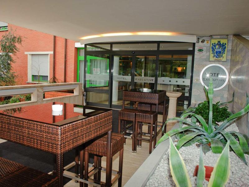 Überdachte Terrasse mit braunen Holztischen und Stühlen, Pflanzen und Glasvitrine im Hintergrund.