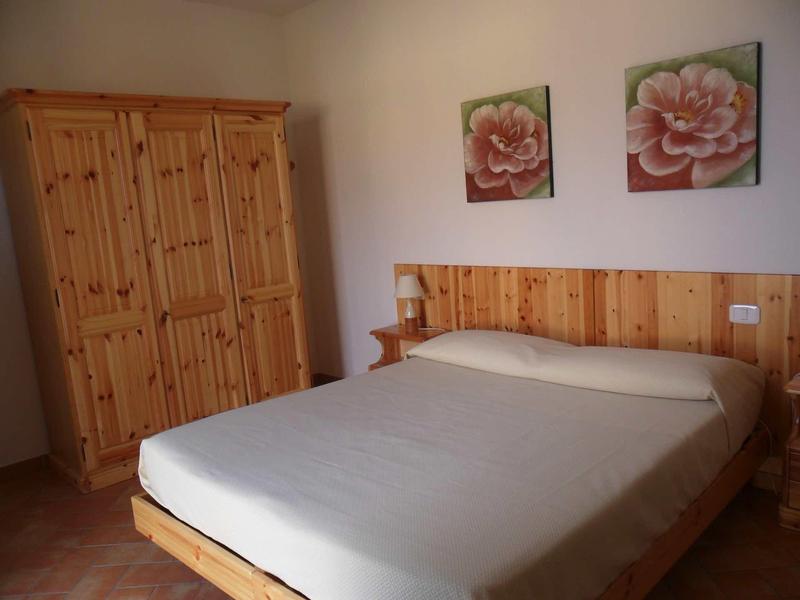 Schlafzimmer mit Holzschrank, Doppelbett mit hellen Bettlaken und zwei Blumenbildern an der Wand.
