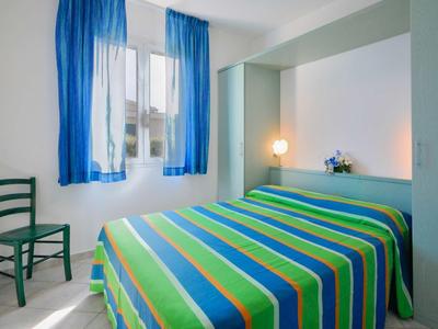 Kleines Schlafzimmer mit grün-blau gestreiftem Bett, blauem Fenstervorhang und grünem Stuhl.