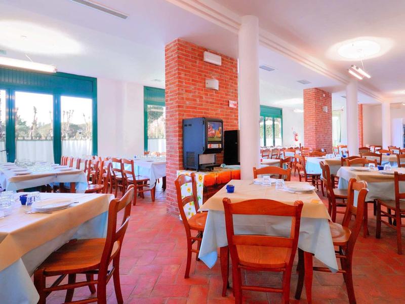 Helles Restaurant mit vielen Holztischen und Stühlen, weißen Tischdecken und großen Fenstern.