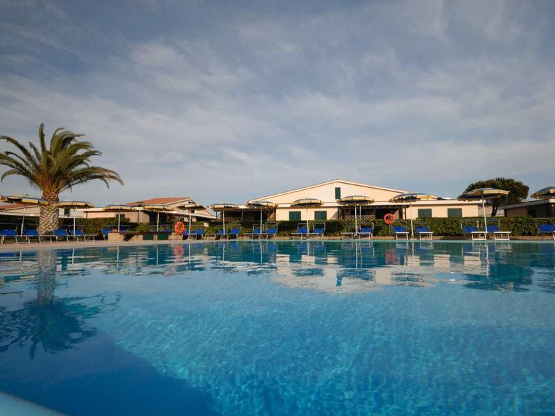 Großer Pool mit blauem Wasser, Sonnenschirme, Palmen und ein Gebäude im Hintergrund bei bewölktem Himmel.