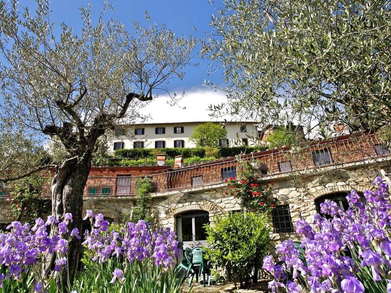 Großes Haus auf Hang mit blühenden violetten Blumen und alten Bäumen unter blauem Himmel.
