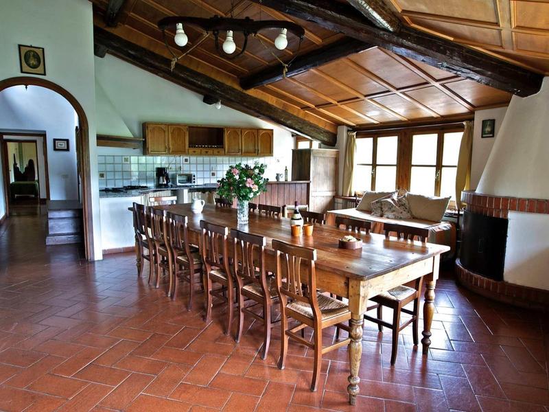 Großer Essbereich mit langem Holztisch, vielen Stühlen, Kaminecke und offener Küche mit Holzbalkendecke.