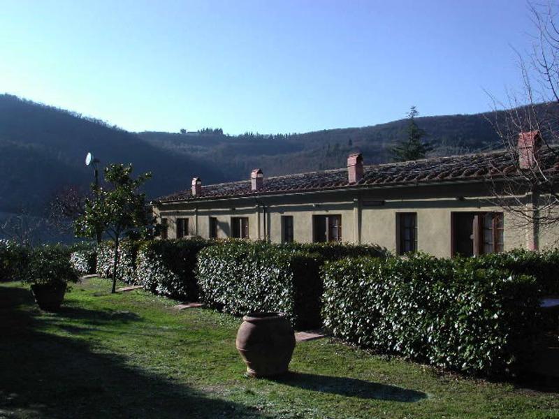 Ländliches Gebäude mit Dachziegeln, davor grüne Hecken, Grasfläche und ein großer Blumentopf.