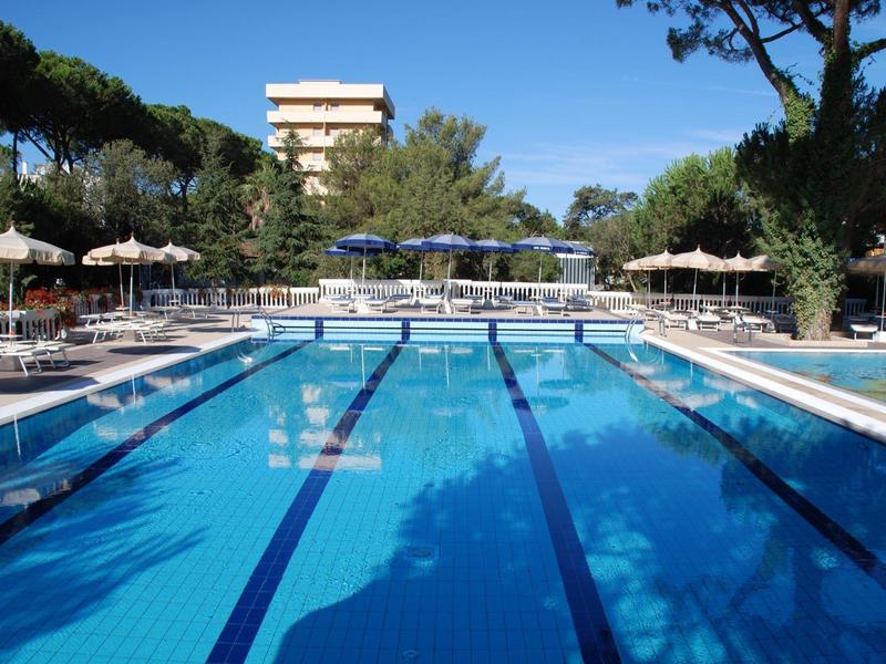 Large outdoor pool with lounge chairs and umbrellas surrounded by trees and buildings.