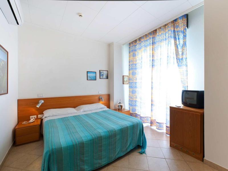 Een eenvoudige hotelkamer met een tweepersoonsbed, klein nachtkastje, tv en raam met lichte gordijnen.