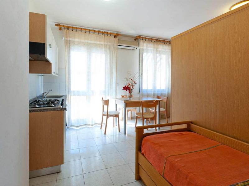 Kleine hotelkamer met kitchenette, eettafel en eenpersoonsbed met oranje bedsprei.