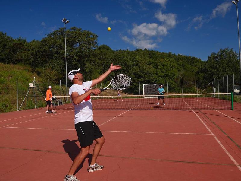 Tennisspieler in weißem Shirt und schwarzer Hose auf rotem Platz bei sonnigem Himmel.