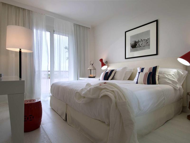 Helles Schlafzimmer mit großem Bett, weißen Wänden, roter Deko und Fenster mit Gardinen.