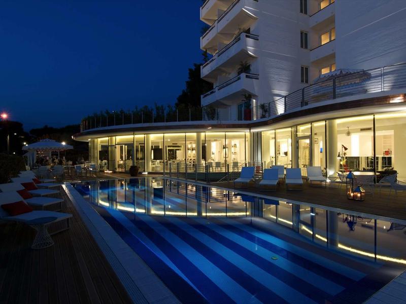 Modernes Hotel mit beleuchtetem Außenpool und Liegestühlen bei Abenddämmerung.